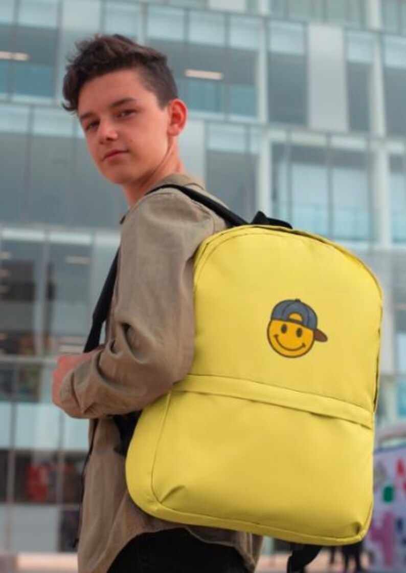 May include: A yellow backpack with a smiling face wearing a baseball cap. The backpack has a front pocket and adjustable straps.