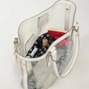 Pu&ograve; includere: Una borsa tote trasparente in plastica con un bordo bianco. La borsa &egrave; aperta e contiene diversi oggetti, tra cui una sciarpa a fantasia, un portafoglio rosso e un oggetto a righe rosa e bianco.