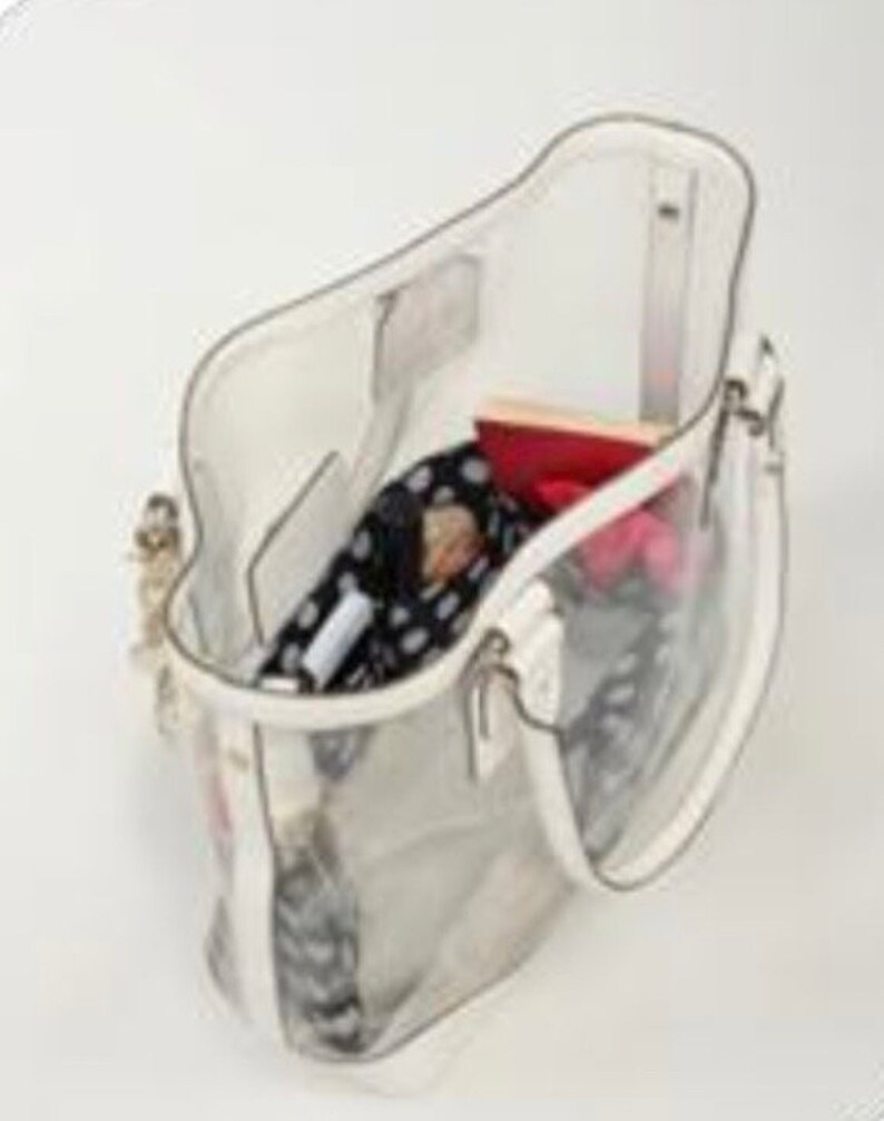 Pu&ograve; includere: Una borsa tote trasparente in plastica con un bordo bianco. La borsa &egrave; aperta e contiene diversi oggetti, tra cui una sciarpa a fantasia, un portafoglio rosso e un oggetto a righe rosa e bianco.