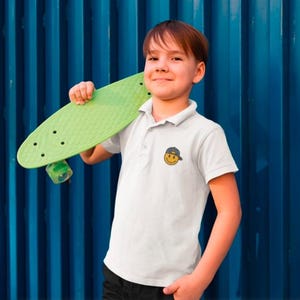 May include: A young person wearing a white polo shirt with a yellow smiley face graphic wearing a blue baseball cap. The person is holding a green skateboard.