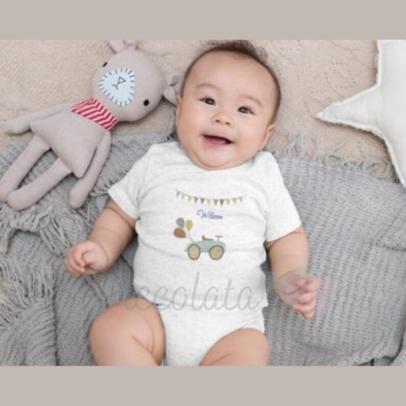 May include: A white baby onesie with a blue and gray toy truck and balloons on the front. The onesie has a banner with the name "William" on it.