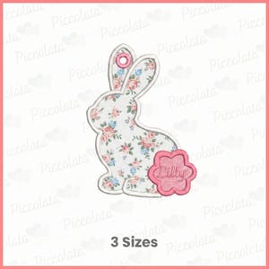 Könnte beinhalten: Ein blumenförmiges Hasen-Applikation mit einem rosa blütenförmigen Anhänger mit der Aufschrift "Lilly". Der Hase hat einen weißen Hintergrund mit einem Blumenmuster aus rosa und blauen Blumen. Der Text "3 Sizes" steht unten.
