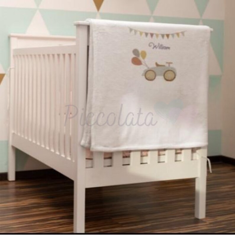 May include: White crib with a white and gray personalized baby blanket. The blanket has a small car with balloons and the name "William" embroidered on it.