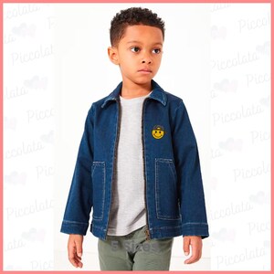 May include: A young boy wearing a blue denim jacket with a yellow smiley face patch. The jacket has a zipper closure and two pockets on the front. The boy is wearing a grey t-shirt and green pants.