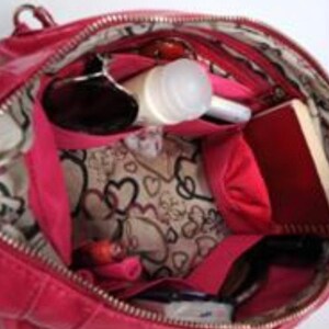 Pu&ograve; includere: Una borsa a mano in pelle rosa con chiusura a zip e fodera a fantasia bianca e rosa. La borsa &egrave; aperta e piena di vari oggetti, tra cui occhiali da sole, una bottiglia bianca, un taccuino rosa e una trousse da trucco rosa.