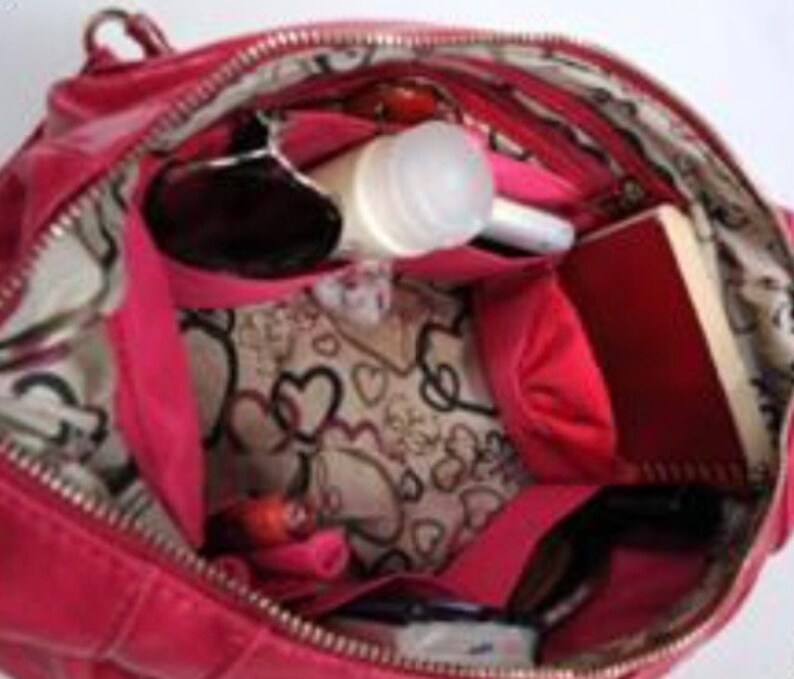 Pu&ograve; includere: Una borsa a mano in pelle rosa con chiusura a zip e fodera a fantasia bianca e rosa. La borsa &egrave; aperta e piena di vari oggetti, tra cui occhiali da sole, una bottiglia bianca, un taccuino rosa e una trousse da trucco rosa.
