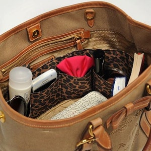 Pu&ograve; includere: L'interno di una borsa tote in tela marrone con rifiniture in pelle marrone. La borsa &egrave; foderata con un tessuto a stampa leopardata e ha diverse tasche. Le tasche contengono un telefono cellulare, un fazzoletto rosso, una bottiglia di profumo, un libro e altri oggetti.