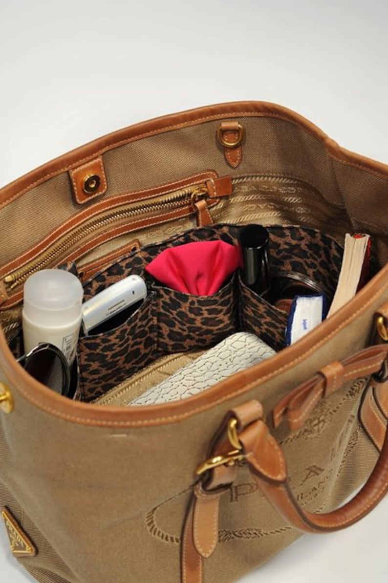 Pu&ograve; includere: L'interno di una borsa tote in tela marrone con rifiniture in pelle marrone. La borsa &egrave; foderata con un tessuto a stampa leopardata e ha diverse tasche. Le tasche contengono un telefono cellulare, un fazzoletto rosso, una bottiglia di profumo, un libro e altri oggetti.