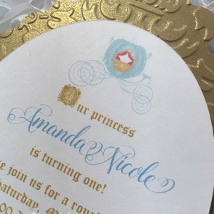 May include: A white and gold invitation with a blue and gold Cinderella carriage design. The invitation reads "Our princess Amanda Nicole is turning one!" and includes the text "Join us for a royal celebration Saturday, October 25th at noon.  Keepsakes by Ingrid 813.778.2700 www.keepsakesbyingrid.weebly.com"