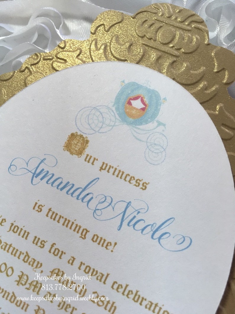 May include: A white and gold invitation with a blue and gold Cinderella carriage design. The invitation reads "Our princess Amanda Nicole is turning one!" and includes the text "Join us for a royal celebration Saturday, October 25th at noon.  Keepsakes by Ingrid 813.778.2700 www.keepsakesbyingrid.weebly.com"