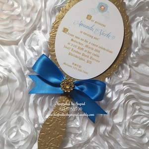 May include: Gold mirror-shaped invitation with a blue ribbon and a gold bow. The invitation is for a princess's first birthday party. The text on the invitation reads "Our princess Amanda Nicole is turning one! Please join us for a royal celebration Saturday, March 28th at 3:00 PM at her castle 5447 Lyndon Park Terrace Philadelphia, PA RSVP: Melanie 123.456.7890 Keepsakes by Ingrid 813.778.2700 www.keepsakesbyingrid.weebly.com"