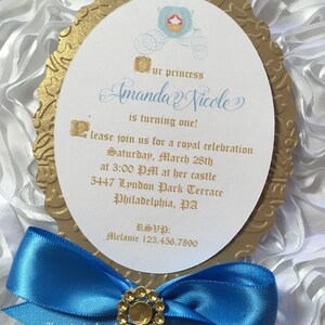 May include: A gold-colored mirror-shaped invitation with a blue ribbon and a rhinestone embellishment. The invitation is for a princess-themed first birthday party for Amanda Nicole. The invitation includes the date, time, and location of the party.