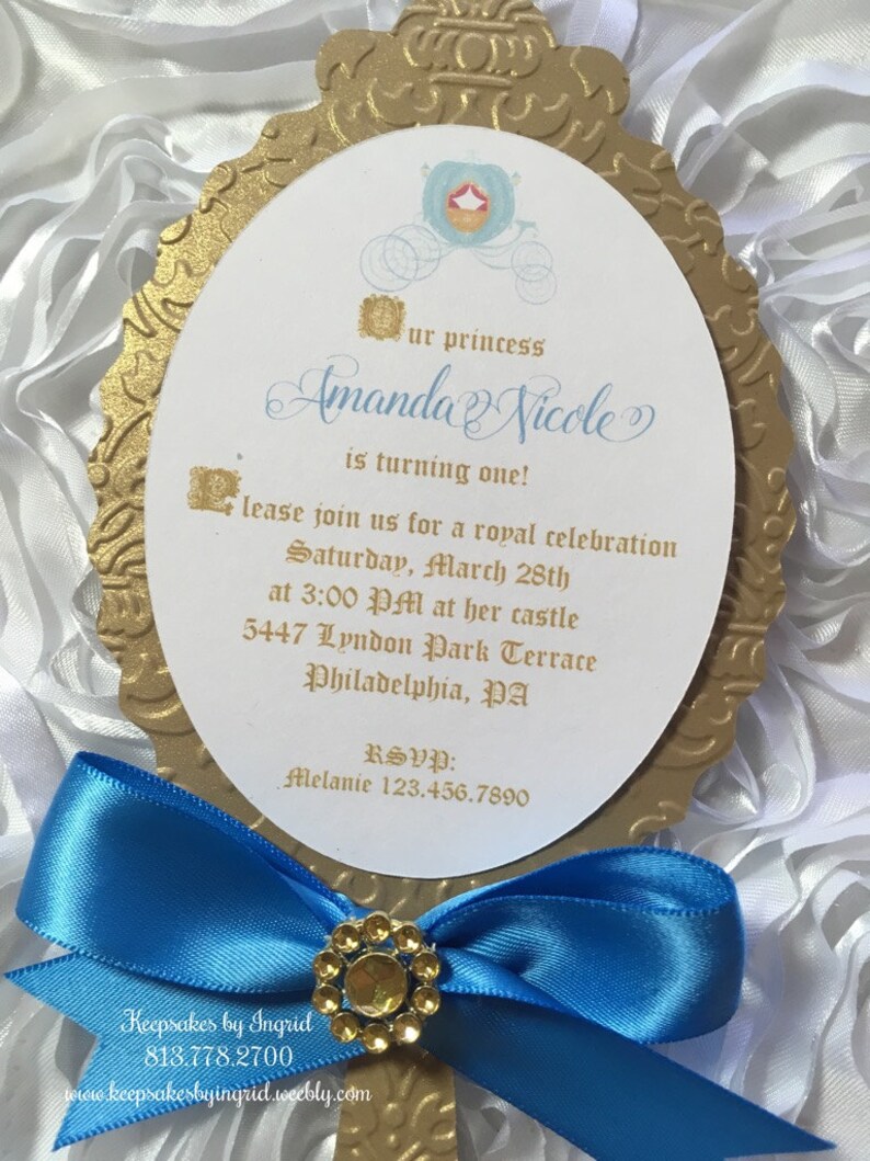 May include: A gold-colored mirror-shaped invitation with a blue ribbon and a rhinestone embellishment. The invitation is for a princess-themed first birthday party for Amanda Nicole. The invitation includes the date, time, and location of the party.