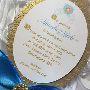 May include: Gold and blue invitation with a Cinderella carriage design. The invitation reads "Our princess Amanda Nicole is turning one!" and includes details for a royal celebration on Saturday, March 28th at 3:00 PM. The address is 5447 Lyndon Park Terrace, Philadelphia, PA. RSVP: Melanie 123.456.7890.
