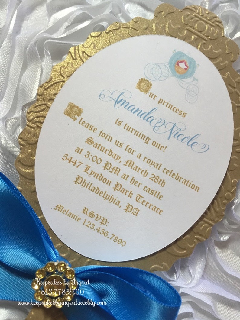 May include: Gold and blue invitation with a Cinderella carriage design. The invitation reads "Our princess Amanda Nicole is turning one!" and includes details for a royal celebration on Saturday, March 28th at 3:00 PM. The address is 5447 Lyndon Park Terrace, Philadelphia, PA. RSVP: Melanie 123.456.7890.