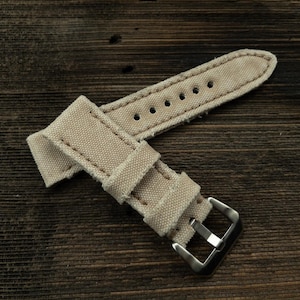Könnte beinhalten: Ein beigefarbenes Canvas-Armband mit einer silbernen Schließe. Das Armband ist in der Mitte gefaltet und die Schließe ist sichtbar.