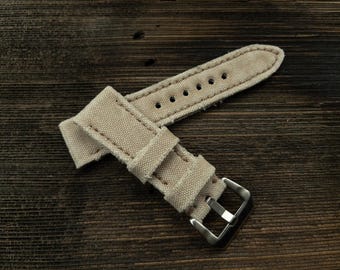 May include: A beige canvas watch strap with a silver buckle. The strap is folded in half and the buckle is visible.