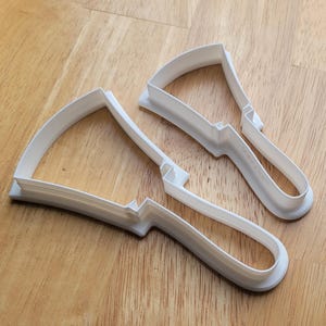 May include: Two white plastic cookie cutters in the shape of a wedge of cheese with a handle. The cutters are on a wooden surface. The cutters are designed to cut out the shape of a wedge of cheese from cookie dough.