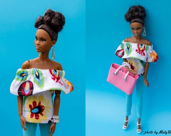 May include: A doll wearing a white off-the-shoulder top with a colorful floral pattern, blue jeans, and a pink handbag. The doll has dark brown hair and brown eyes.