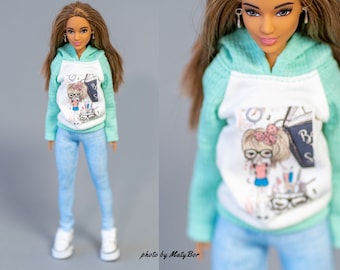 May include: A doll wearing a light blue and white hooded sweatshirt with a graphic print of a girl wearing glasses and holding a book. The doll is also wearing light blue jeans and white sneakers.