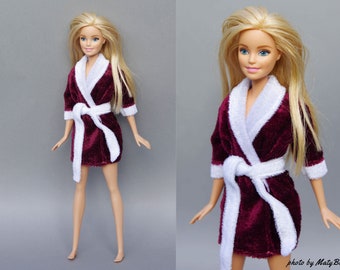 Może przedstawiać: Lalka Barbie w burgundowej szlafroku z aksamitu z białymi lamówkami i białym pasem. Szlafrok ma kołnierz szalowy i długie rękawy.