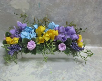 May include: A white planter box filled with a floral arrangement of purple, blue, yellow, and pink flowers. The flowers are arranged in a cascading style, with the purple flowers at the top and the pink flowers at the bottom.