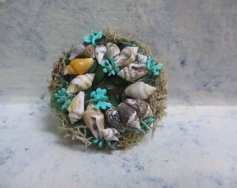 May include: A wreath made of moss and seashells with turquoise flowers. The wreath is decorated with a variety of seashells, including some with a yellow and brown pattern.