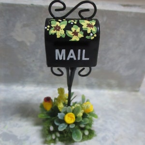 May include: Black metal mailbox with yellow flowers painted on the front. The mailbox is on a stand with a small flower arrangement at the base.