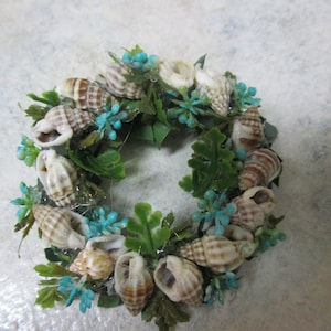 May include: A small wreath made of seashells and faux greenery. The wreath is decorated with blue flowers and has a rustic, coastal feel.