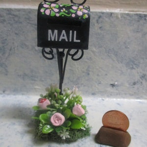 May include: A miniature black mailbox with pink and green flowers painted on it. The mailbox is attached to a black metal stand with a decorative scroll design. The mailbox is surrounded by a small bed of artificial flowers and greenery. A copper penny is sitting on a small brown base.