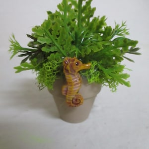 May include: A small, beige, tapered pot containing a cluster of vibrant green artificial foliage. A miniature, textured, yellow and orange seahorse figurine is attached to the front rim of the pot, facing toward the right side of the frame.