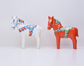 Puede incluir: Dos figuritas de caballos de madera, una blanca con detalles pintados en azul y rojo y otra roja con detalles pintados en azul y verde.