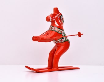 Puede incluir: Una figurita de caballo Dala de madera roja en esquís, sosteniendo bastones de esquí. El caballo está pintado con diseños tradicionales de arte popular sueco en verde, blanco y negro.