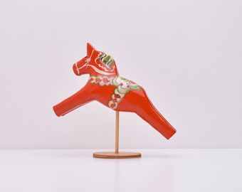 Puede incluir: Un caballo de madera rojo con detalles pintados, conocido como caballo Dala, está sobre un soporte de madera. El caballo está pintado con diseños florales blancos y verdes.