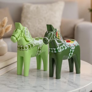 Op de afbeelding: Twee houten paardenbeeldjes met geschilderde bloemmotieven. Het ene paard is lichtgroen met witte details, terwijl het andere donker bosgroen is met witte, gele en rode accenten. Beide hebben gestileerde manen en zadels.
