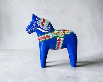 May include: A blue wooden horse figurine featuring hand-painted floral and geometric patterns in red, yellow, green, and white. The horse stands in profile, displaying a glossy finish with intricate decorative details on its mane, back, and sides.