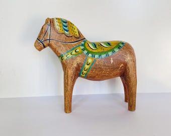 Puede incluir: Figura de caballo Dala de madera pintada a mano. El caballo es marrón con detalles pintados de colores amarillo, verde y azul. El caballo tiene una silla de montar y una brida. La figura es un objeto de arte popular sueco tradicional.