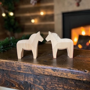 Pode incluir: Duas figuras de cavalos em madeira de cor clara esculpidas com linhas suaves e angulares. Cada cavalo apoia-se em quatro patas com um pescoço curvo e um acabamento simples sem pintura que realça o grão natural da madeira clara.
