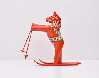 Puede incluir: Una figurita de caballo Dala de madera roja en esquís con detalles florales pintados. El caballo sostiene bastones de esquí en sus patas delanteras.