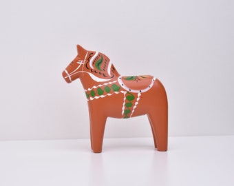 Könnte beinhalten: Eine traditionelle schwedische Dala-Pferdefigur, in klassischem Rot mit weißen und grünen Details bemalt. Das Pferd hat einen Sattel und ein Zaumzeug und ist ein beliebtes skandinavisches Volkskunststück.