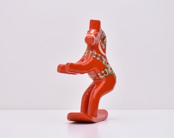 May include: A vibrant red Dala horse figurine with a white and patterned design on its head and body. The wooden horse has a curved body and is standing on a flat base. A traditional Swedish folk art item.