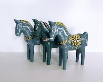 May include: Three hand-painted wooden horse figurines in shades of blue and green. Each horse has a yellow saddle and mane, with white accents. The horses stand side-by-side, showcasing a folk art style.