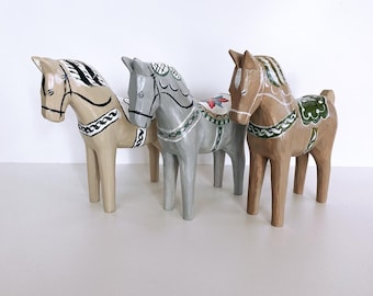 Könnte beinhalten: Drei Holzpferde-Figuren, in verschiedenen Farben bemalt. Die Pferde stehen auf einer weißen Oberfläche. Das Pferd links ist beige mit schwarzen und weißen Details. Das Pferd in der Mitte ist grau mit schwarzen und grünen Details. Das Pferd rechts ist braun mit schwarzen, grünen und roten Details.