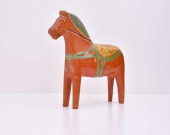Puede incluir: Un caballo Dala sueco tradicional, un juguete de madera en forma de caballo pintado de rojo, verde y amarillo. El caballo tiene una silla de montar y una brida.