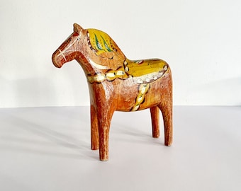 Puede incluir: Una figurita de caballo de madera pintada a mano con detalles amarillos, marrones y verdes. El caballo está de pie sobre una superficie blanca.