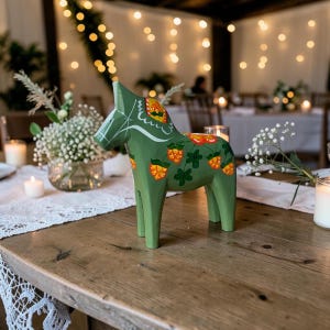 Pode incluir: Uma estatueta de cavalo Dala de madeira em verde sálvia com motivos de amoras-brancas em laranja e amarelo pintados à mão com folhas verdes. O cavalo tem detalhes de arreios pintados em branco e está sobre uma mesa de madeira.