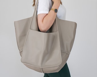 Könnte beinhalten: Eine große, taupefarbene Ledertasche mit einer lockeren, unstrukturierten Silhouette. Die Tasche verfügt über einen breiten Schulterriemen, eine markante Vordertasche mit sichtbaren Nähten und eine glatte, matte Oberfläche auf der Außenseite.