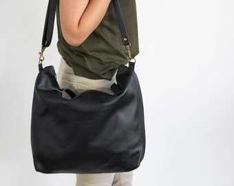 May include: A black leather hobo bag with a slouchy silhouette, featuring a wide shoulder strap attached with antique brass-toned metal swivel clips. The bag has a smooth, textured surface and a simple, unadorned design with a top zipper closure.