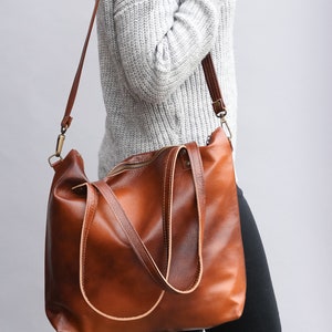 May include: A large, cognac-brown leather tote bag featuring a smooth, polished finish, a top zipper closure, and two sturdy shoulder straps. The bag includes a detachable, adjustable long crossbody strap with antique brass-toned metal hardware.