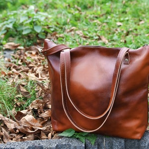 May include: A medium-sized brown leather tote bag with a smooth, polished finish. The bag features two long, thin shoulder straps, a top zipper closure, and metallic hardware attachments on the sides for an adjustable strap.